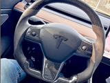 Steering wheel with a carbon fiber design displays a Tesla logo, held by a person wearing blue jeans inside a car. A wooden dashboard and vehicle interior surround it.