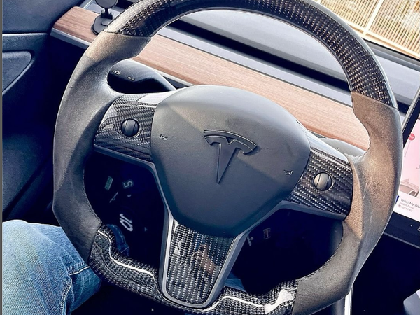 Steering wheel with a carbon fiber design displays a Tesla logo, held by a person wearing blue jeans inside a car. A wooden dashboard and vehicle interior surround it.
