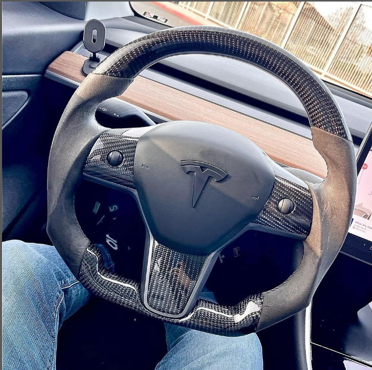 Steering wheel with a carbon fiber design displays a Tesla logo, held by a person wearing blue jeans inside a car. A wooden dashboard and vehicle interior surround it.