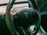 A steering wheel with a Tesla logo is mounted in a car's interior, featuring a touchscreen dashboard and luxurious black detailing. The environment suggests a modern, sophisticated vehicle interior.