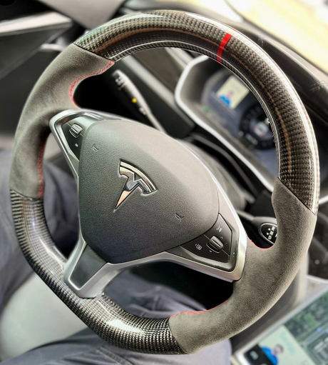 Steering wheel with a carbon fiber and suede design, featuring a metallic logo and control buttons; situated in a modern car interior with digital display screens visible.