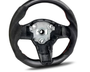 A carbon fiber steering wheel with a central metal hub is positioned upright. It features red stitching and lacks controls, set against a plain white background.