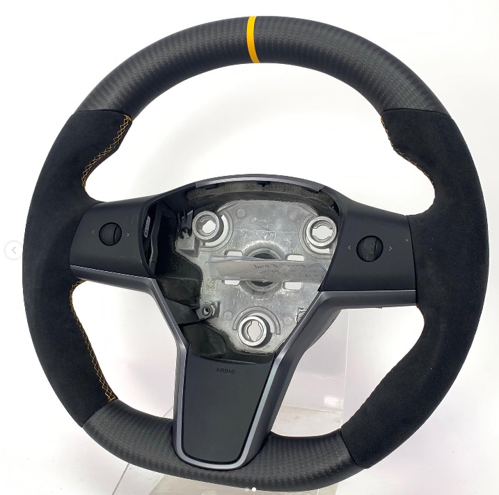 A racing steering wheel with a carbon fiber and suede grip features a yellow top centering stripe. It's positioned against a plain white background, showing exposed mounting components.