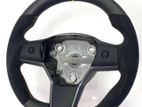 A steering wheel with a central mounting plate and integrated controls sits against a plain white background. The wheel has a black suede grip with a yellow top marker and detailed stitching.