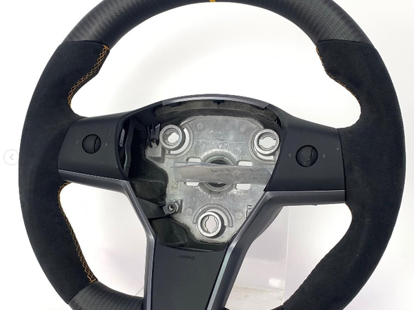 A steering wheel with a central mounting plate and integrated controls sits against a plain white background. The wheel has a black suede grip with a yellow top marker and detailed stitching.