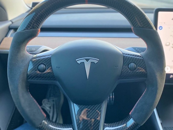 Steering wheel with a carbon fiber finish and logo, held by unseen hands. It is inside a car with a digital display to the right, legs visible below.