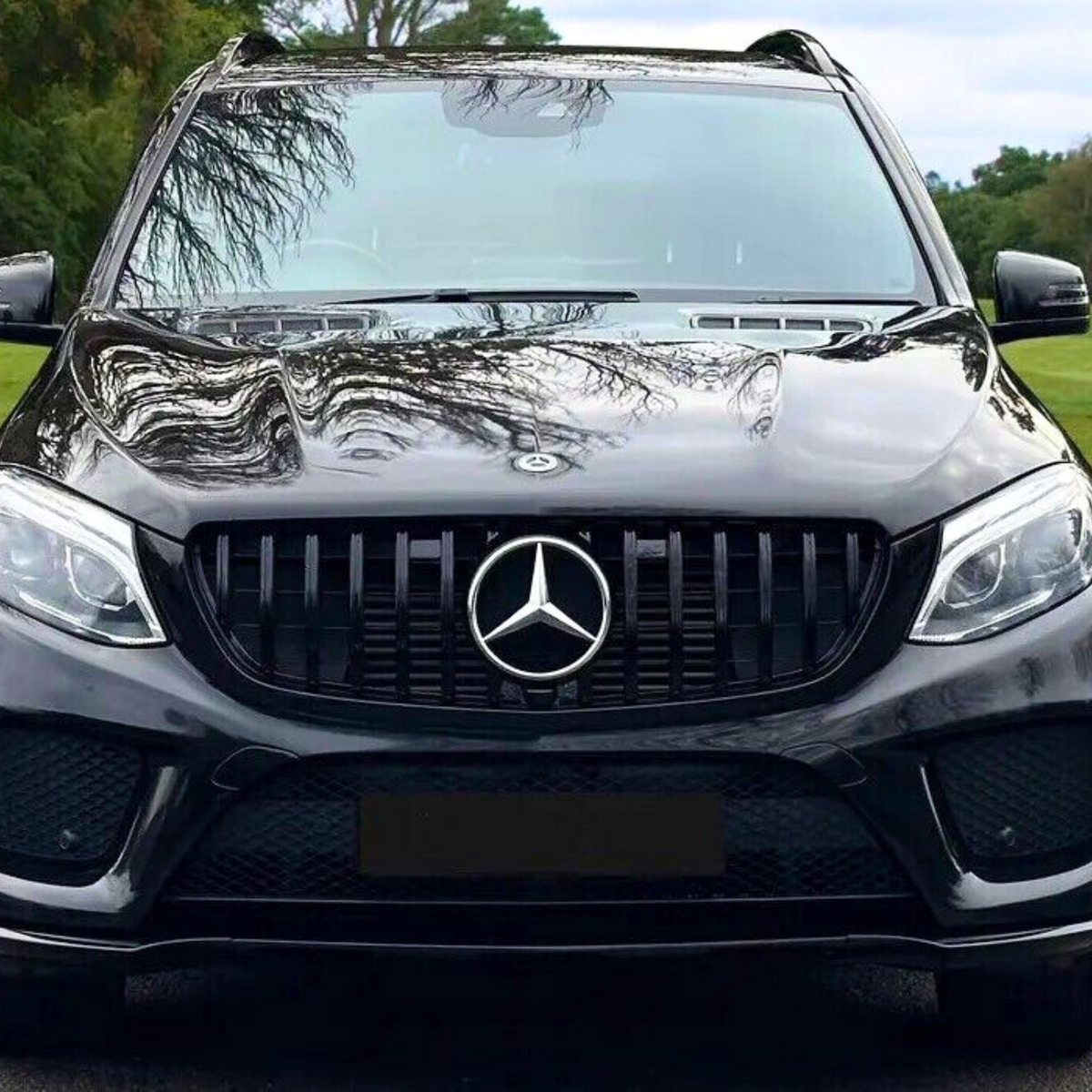 A black SUV is parked on a grassy area, showing its front with a prominent Mercedes emblem on the grille, surrounded by trees in the background.