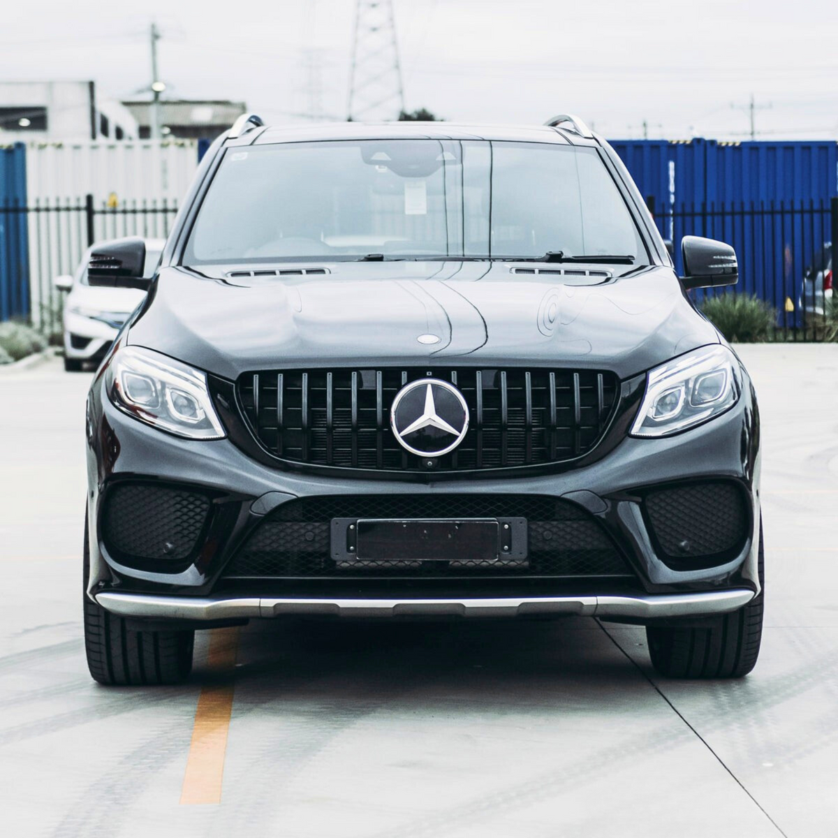 A black Mercedes SUV is parked on a concrete surface. The background features a blue container, fencing, and industrial buildings under an overcast sky.
