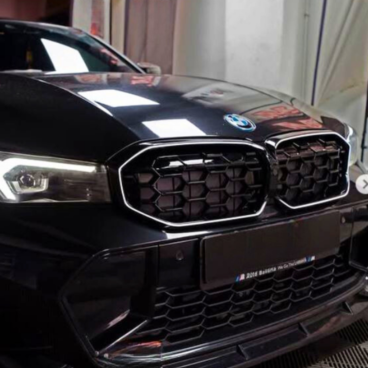 A black car's front, featuring a distinctive hexagonal grille and illuminated headlights, is parked indoors. Text on the license plate reads: "M 2Auto Bavaria. We Go The Extra Mile."