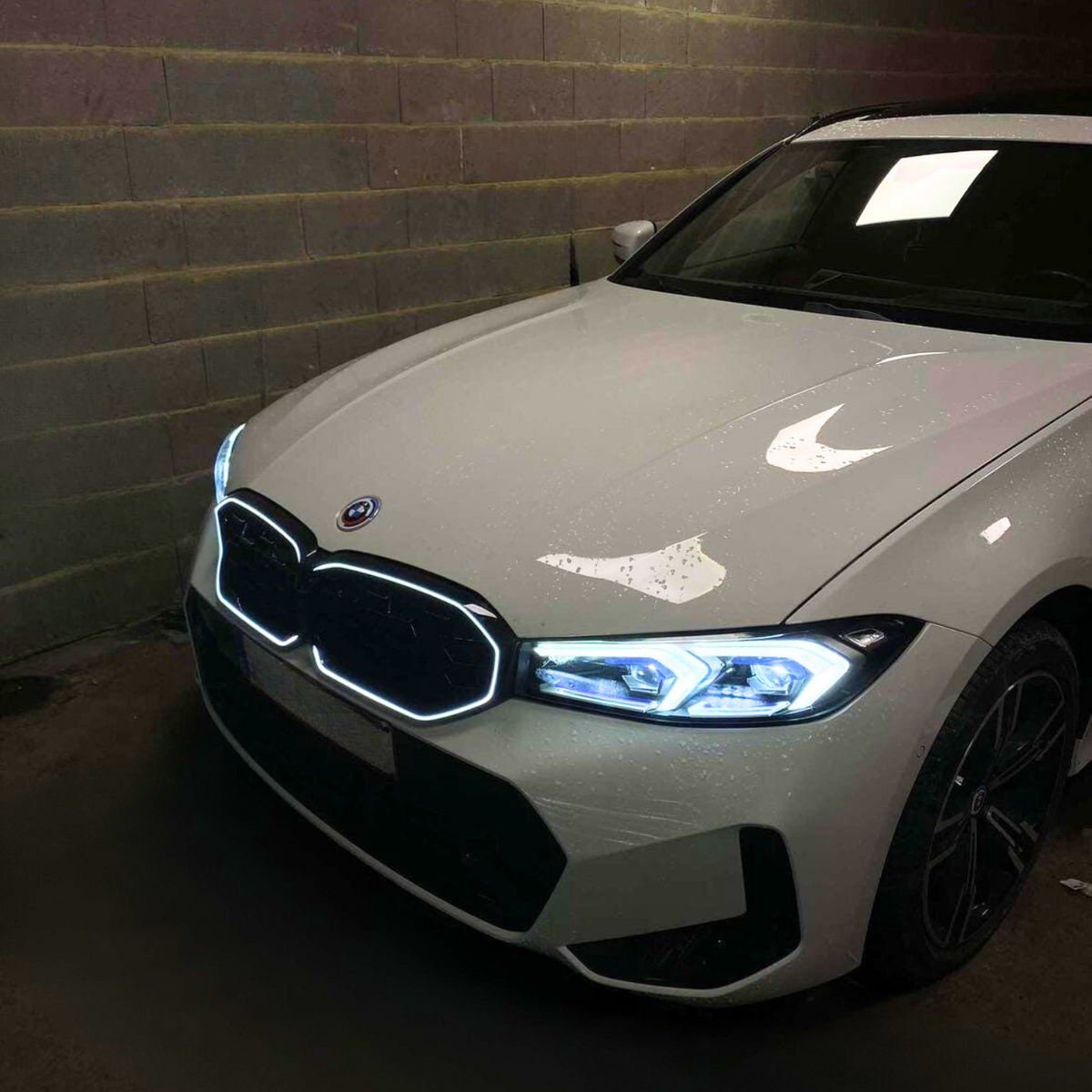 A white car with illuminated headlights parked in a dimly lit garage with brick walls.