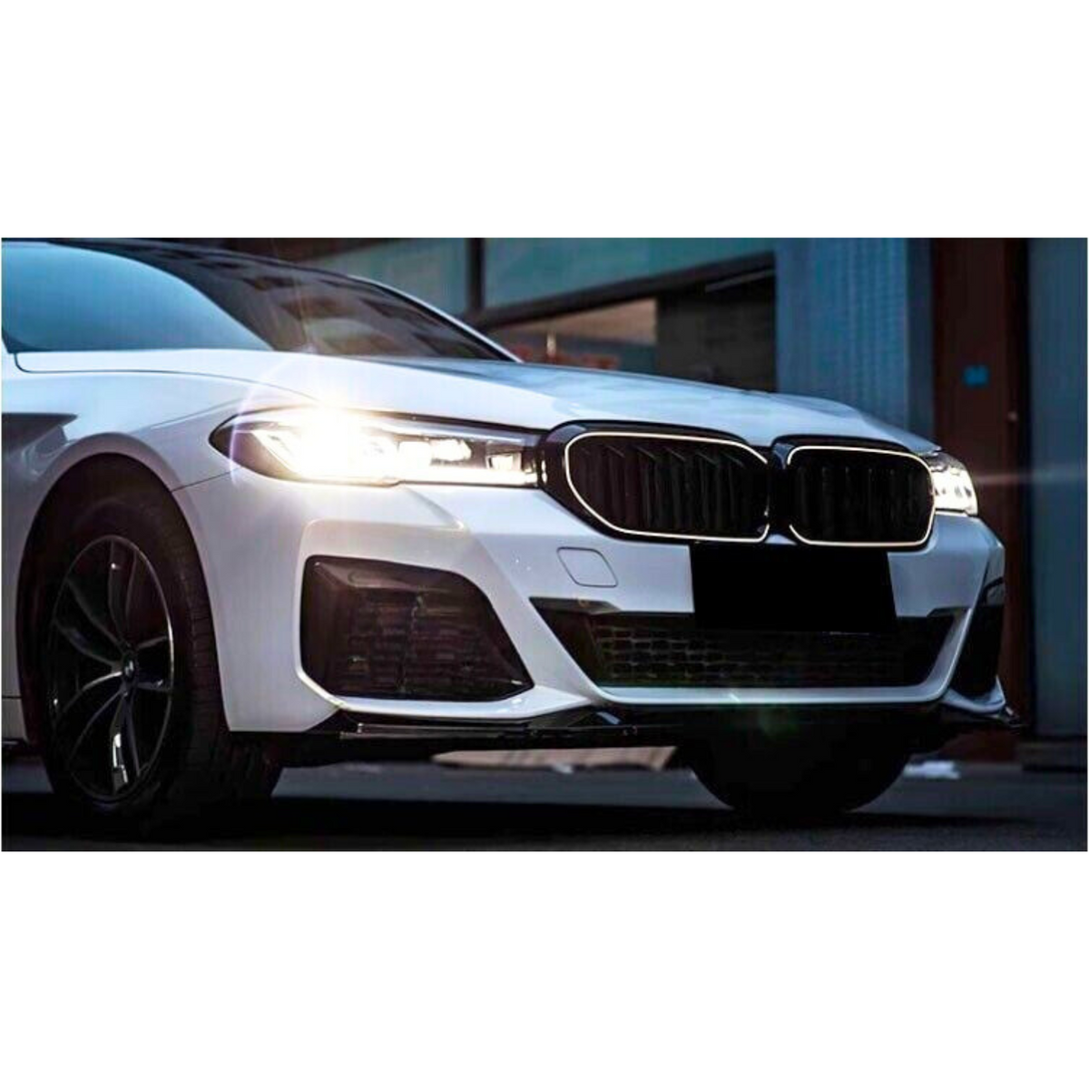 A white car with illuminated headlights is parked on a street, featuring a prominent front grille and sleek design, partially surrounded by urban buildings.