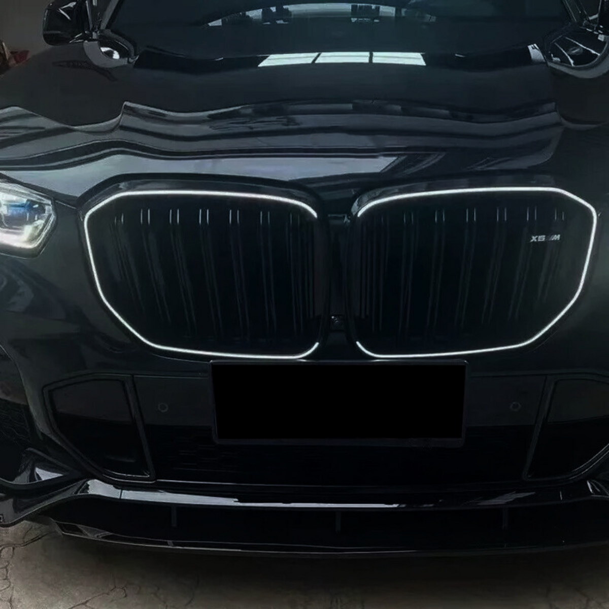 A black car with a prominent, illuminated grille, showcasing a sleek design. The front is parked indoors, with reflections visible on the shiny surface. "X5 M" is marked on the grille.
