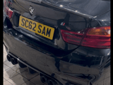 A black BMW car revs its engine, emitting exhaust fumes. The car is parked indoors, showcasing the rear view, with the license plate reading 