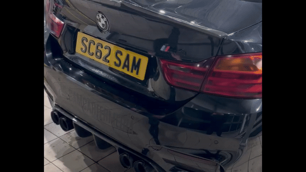 A black BMW car revs its engine, emitting exhaust fumes. The car is parked indoors, showcasing the rear view, with the license plate reading "SC62 SAM" and distinctive red taillights.