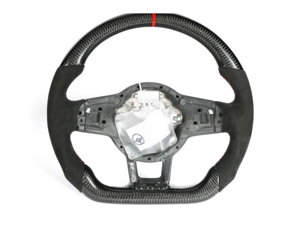 A carbon fiber and suede steering wheel with a red center stripe is displayed against a plain white background. Its center is exposed, revealing inner components, and a small plastic bag is attached.