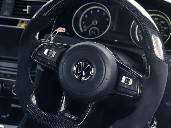 A car steering wheel with a Volkswagen logo, featuring control buttons, is set in a vehicle interior with visible speedometer and climate control dials.