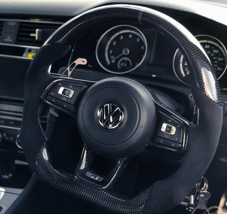 A carbon-fiber steering wheel with a Volkswagen logo is centered in a car's dashboard, featuring control buttons on the spokes and surrounded by visible speedometers and gauges.