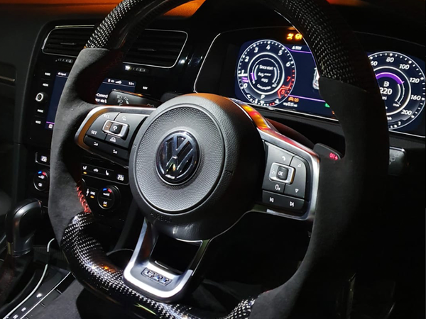 A car steering wheel with a carbon fiber texture is positioned at the center, displaying a Volkswagen logo. Surrounding digital dashboard panels show speedometer and vehicle information in a dimly lit car interior.