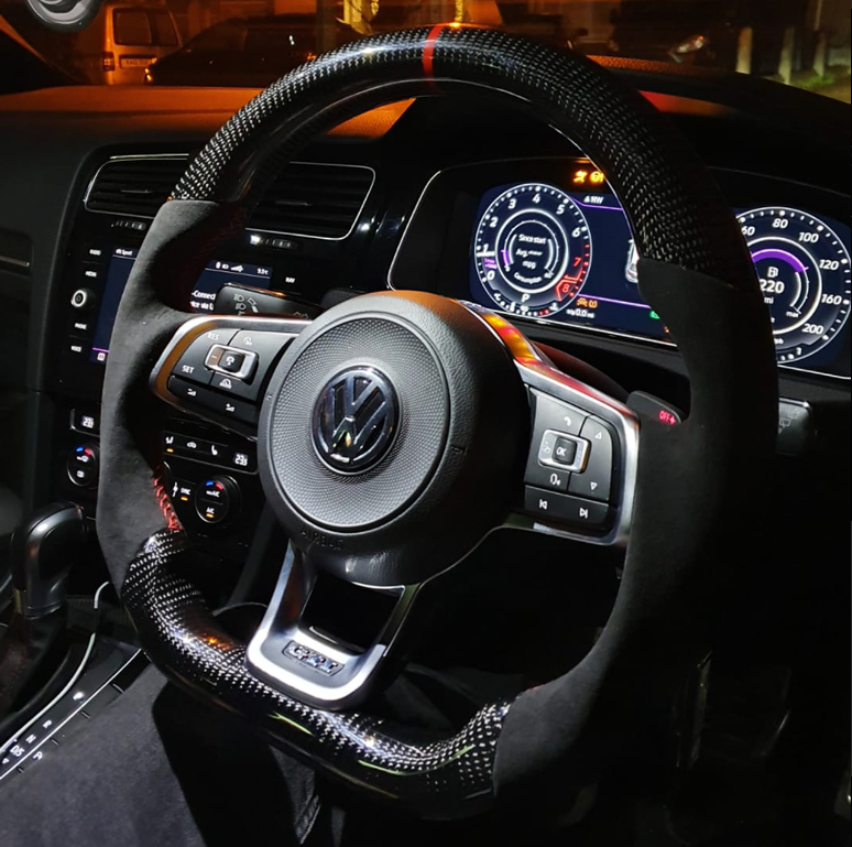 A car steering wheel with a carbon fiber texture is positioned at the center, displaying a Volkswagen logo. Surrounding digital dashboard panels show speedometer and vehicle information in a dimly lit car interior.