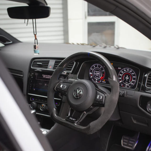Steering wheel with a Volkswagen logo is centered, showing dashboard indicators at 126.0 km/h. Digital displays and controls within a car interior. An air freshener hangs from rearview mirror.