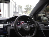 Steering wheel featuring a Volkswagen logo, surrounded by a modern dashboard and digital displays, inside a parked car. The view outside shows a row of buildings and trees.