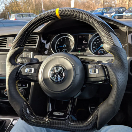 A carbon fiber steering wheel with a yellow stripe is centered in a car's interior, showing buttons and gauges. The background reveals a parking lot with several cars.