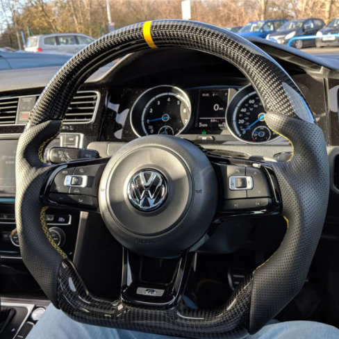 A carbon-fiber steering wheel with a yellow stripe is held by blue-jeaned legs inside a Volkswagen car. The dashboard displays various gauges. Other cars are visible through the windshield.