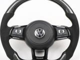 A black steering wheel with a central Volkswagen emblem and multiple control buttons is set against a plain white background. The upper rim features a carbon fiber texture with a red and yellow stripe.