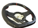 A black carbon fiber car steering wheel with perforated grips and red stitching sits on a white background, displaying its central mounting hub surrounded by intricate details.