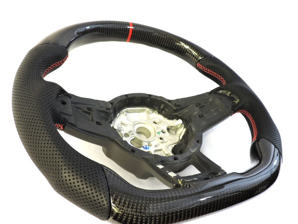 A black carbon fiber car steering wheel with perforated grips and red stitching sits on a white background, displaying its central mounting hub surrounded by intricate details.
