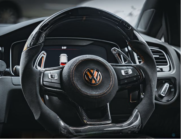 A Volkswagen steering wheel with textured grips, showing controls for various functions, inside a modern car cockpit featuring a digital display and air vent.