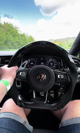Steering wheel gripped, displaying Volkswagen logo, as vehicle travels at 70 mph on a countryside road. Dashboard shows "D6" gear, "2633" odometer, set against a backdrop of trees and clouds.