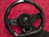 A Volkswagen steering wheel with a chrome and perforated leather finish rests on a red and black textured mat. The central badge displays the VW logo, and 
