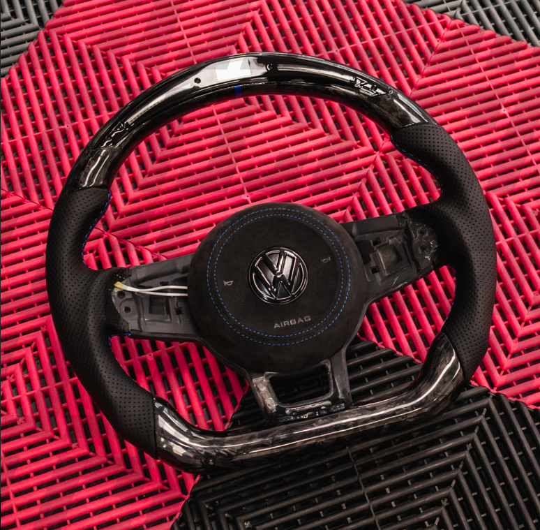 A Volkswagen steering wheel with a chrome and perforated leather finish rests on a red and black textured mat. The central badge displays the VW logo, and "AIRBAG" is labeled beneath it.