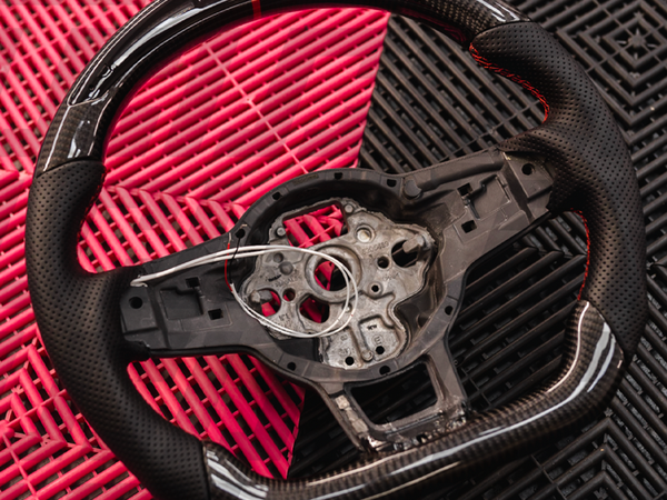 A carbon fiber and black leather steering wheel sits on a red and black textured surface, showing wiring and a circular central area without a cover.