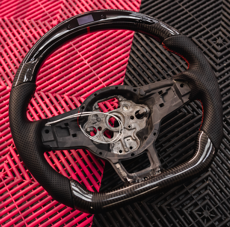 A carbon fiber and black leather steering wheel sits on a red and black textured surface, showing wiring and a circular central area without a cover.