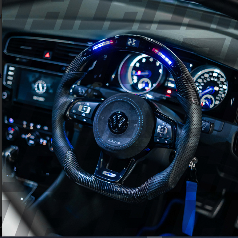 A carbon-fiber steering wheel with a digital display and illuminated strip, mounted in a sleek car dashboard with visible speedometers and controls, indicating a modern automotive interior.