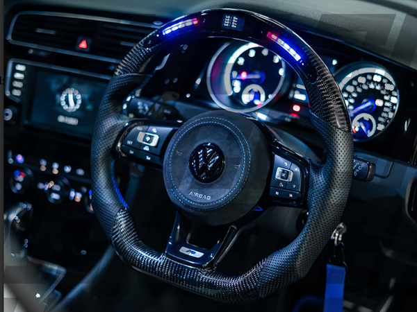 A carbon fiber steering wheel features illuminated controls and a central logo. It is positioned in the dashboard of a car, surrounded by digital speedometers with glowing dials. The word "AIRBAG" is visible.