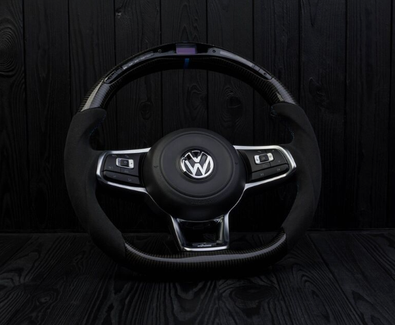 Steering wheel with a Volkswagen logo at the center, featuring integrated buttons; positioned against a black, textured wooden background.