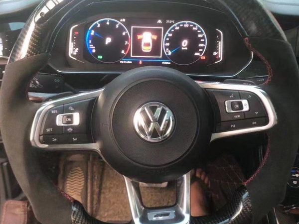 A Volkswagen steering wheel inside a car, featuring control buttons. In the background, a digital dashboard displays driving information with speedometer and gear indicators.