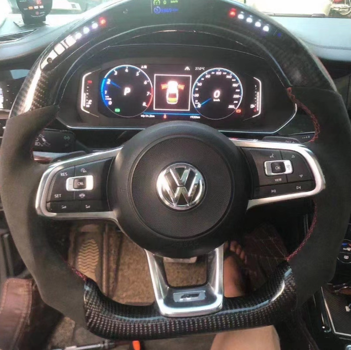 Steering wheel with Volkswagen logo controls a dashboard displaying speedometer, tachometer, and a central digital screen showing a car in "P" mode. Interior features black and carbon fiber accents.