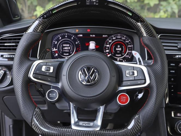 A Volkswagen steering wheel features multifunction controls, including "drive select" and "START STOP" buttons, in a parked car with digital dashboard displaying metrics, surrounded by trees.