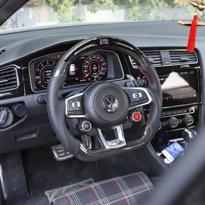 A Volkswagen steering wheel is centered in a car's cockpit, surrounded by digital displays. The dashboard includes various controls and a decorative red tassel hangs from above.
