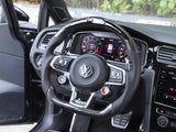 Steering wheel with Volkswagen logo and buttons, positioned in front of a digital dashboard displaying gauges, within a modern car interior featuring a plaid seat.