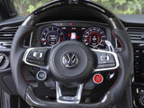 A steering wheel with the Volkswagen logo, featuring buttons and paddle shifters, is centered in a car's interior. The dashboard displays digital gauges, surrounded by lush greenery outside.