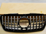 A black car grille with vertical chrome slats and a central circular opening rests against a plain cardboard box on a wooden floor.