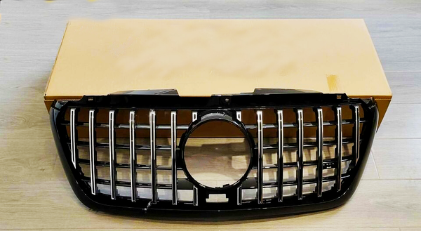 A black car grille with vertical chrome slats and a central circular opening rests against a plain cardboard box on a wooden floor.