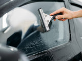 A hand uses a squeegee to wipe water off a car window, with droplets visible on the glass. The setting is a close-up of a car exterior.