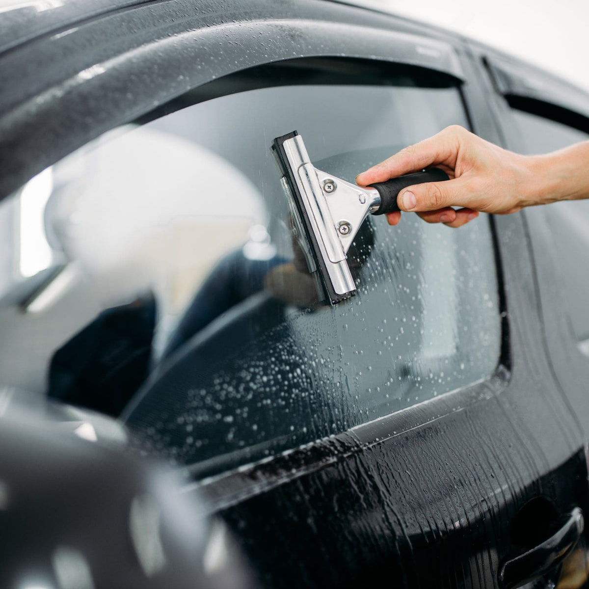 A hand uses a squeegee to wipe water off a car window, with droplets visible on the glass. The setting is a close-up of a car exterior.