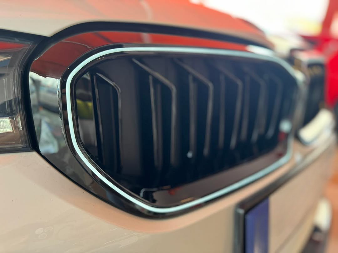 A car's front grille with sleek black vertical slats and shiny chrome trim is positioned in an indoor showroom, with reflections visible on the surface.